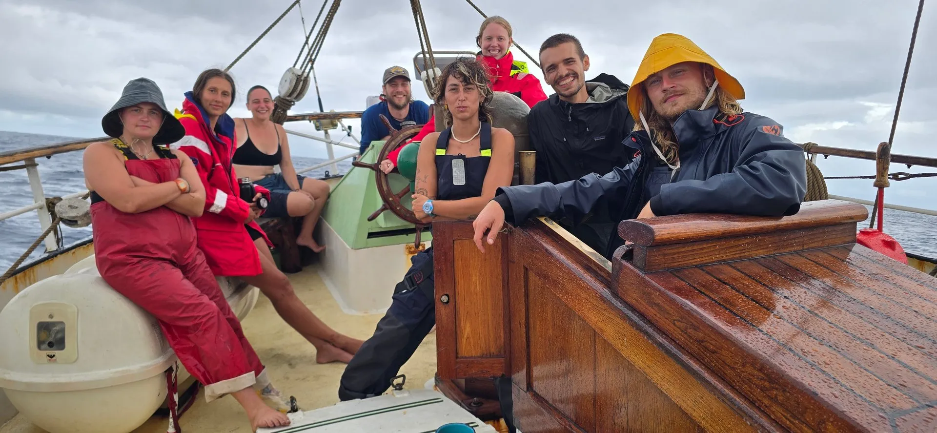 Crew members in wet-weather gear gathered at the helm on a grey day at sea